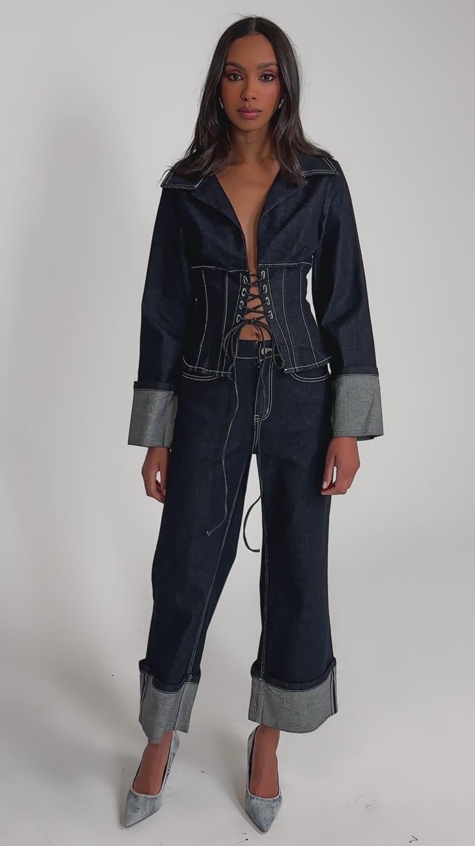 Video of woman modeling the DON'T TALK TO ME LACE UP DENIM BLOUSE, featuring tailored wide cuffed sleeves and a corset-style lace-up front, standing against a plain background with a serious expression.