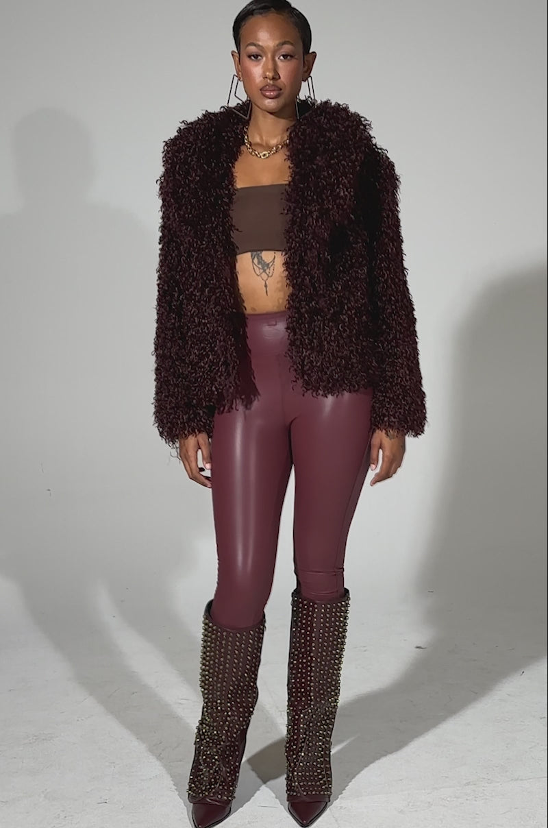 A video of a woman posing against a plain background wearing a textured burgundy faux fur jacket, RIO HIGH RISE LEGGING WITH 4 WAY STRETCH IN OXBLOOD, and tall studded boots. She accessorizes with hoop earrings and has her hair pulled back.