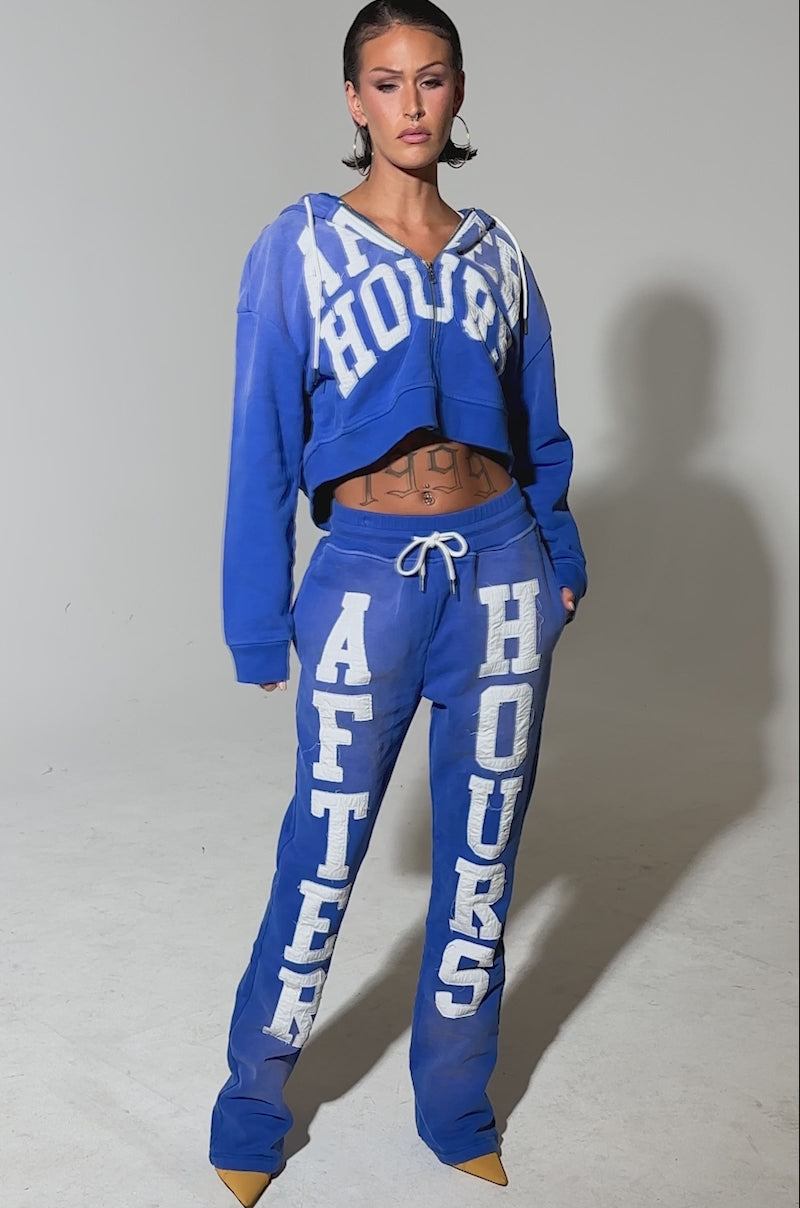 A video of a woman in blue AFTER HOURS FLARED SWEATPANTS with bold white lettering, paired with hoop earrings and yellow pointed shoes, posing against a plain gray background.
