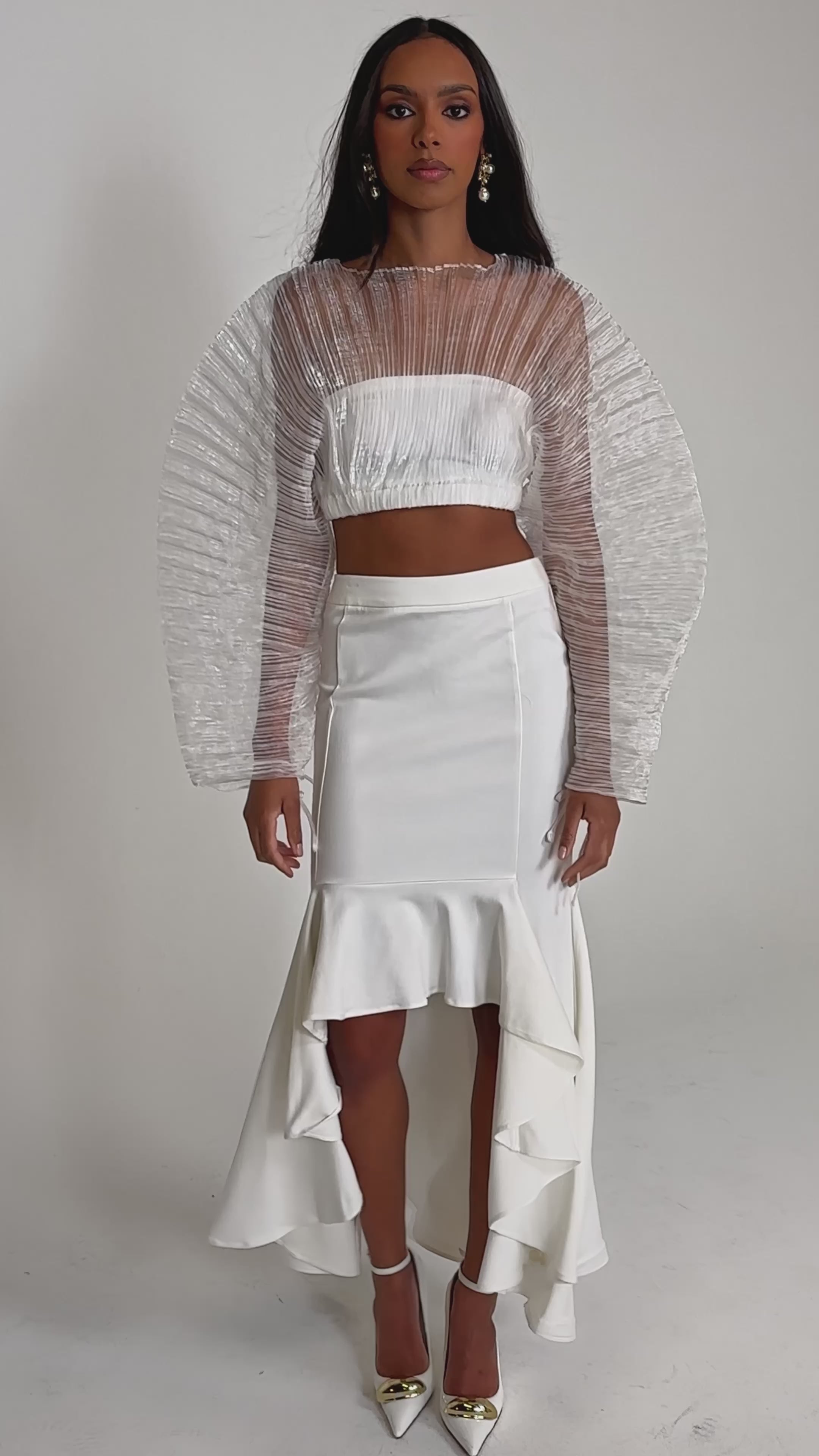 Video of woman modeling the LEMON SATIN MAXI SKIRT with a sheer, textured white long-sleeve crop top and pointed-toe heels, posing against a plain light background.