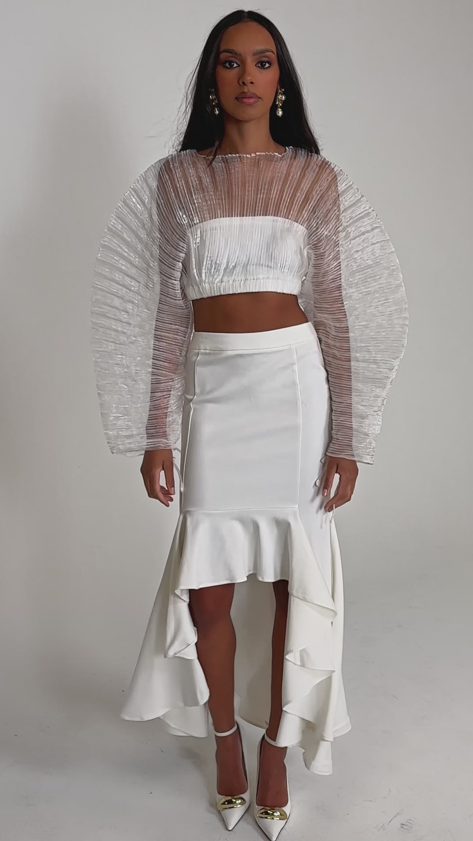 Video of woman modeling the LEMON SATIN MAXI SKIRT with a sheer, textured white long-sleeve crop top and pointed-toe heels, posing against a plain light background.