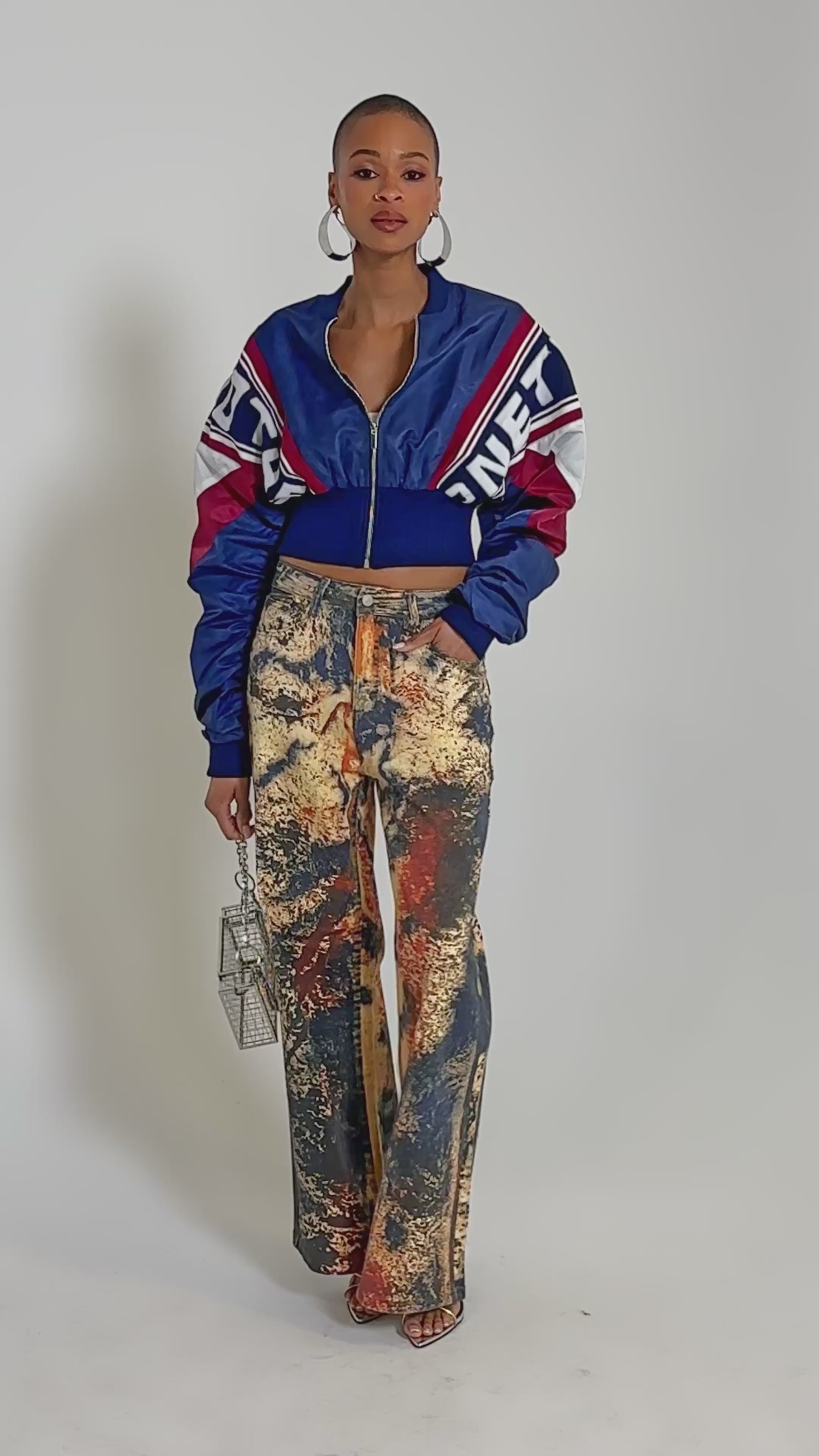 Video of woman modeling the PHOENIX MIXED MEDIA BOMBER IN NAVY, featuring red and white stripes, ruched sleeves, and graphic text details, paired with high-waisted, paint-splattered jeans against a plain light background.