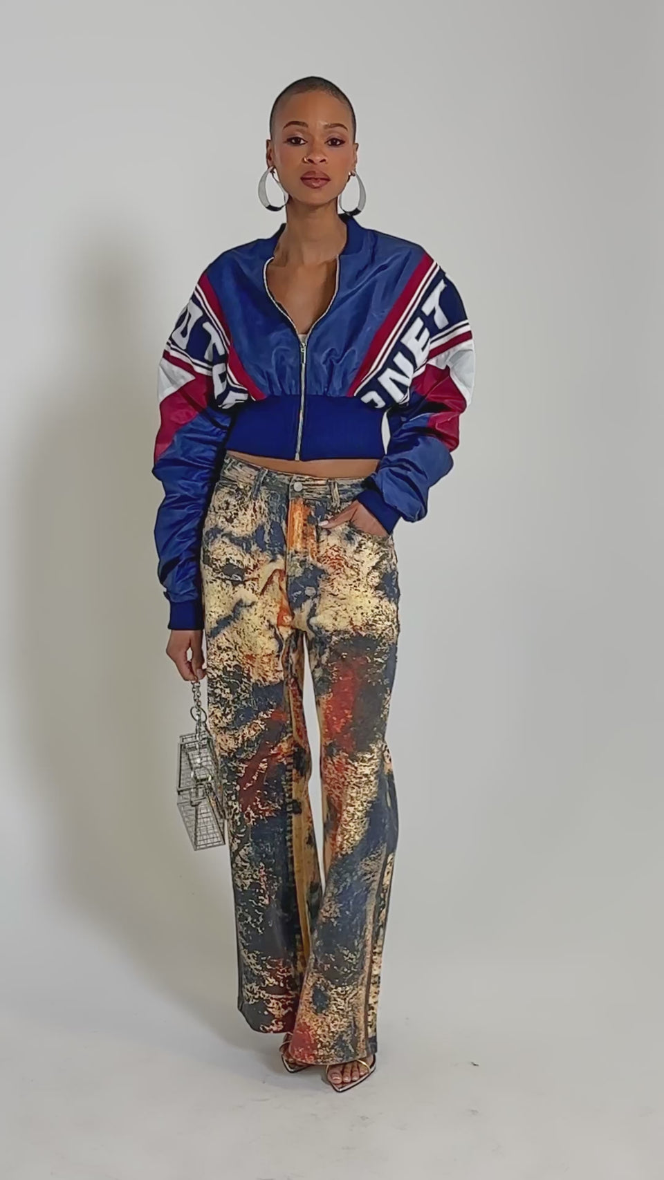 Video of woman modeling the PHOENIX MIXED MEDIA BOMBER IN NAVY, featuring red and white stripes, ruched sleeves, and graphic text details, paired with high-waisted, paint-splattered jeans against a plain light background.