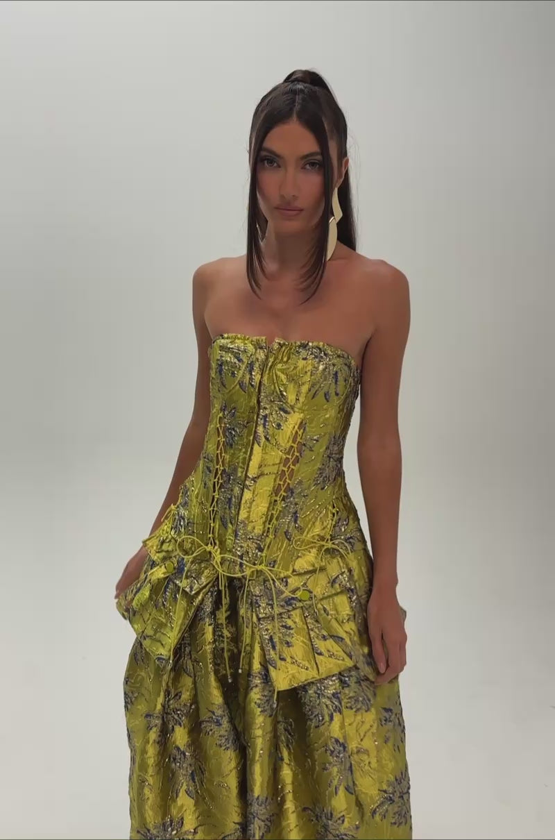 A video of a woman wearing the CRAZY FOR YOU BROCADE CORSET, a strapless metallic green dress with blue floral patterns, lace-up front, and gathered pockets. 