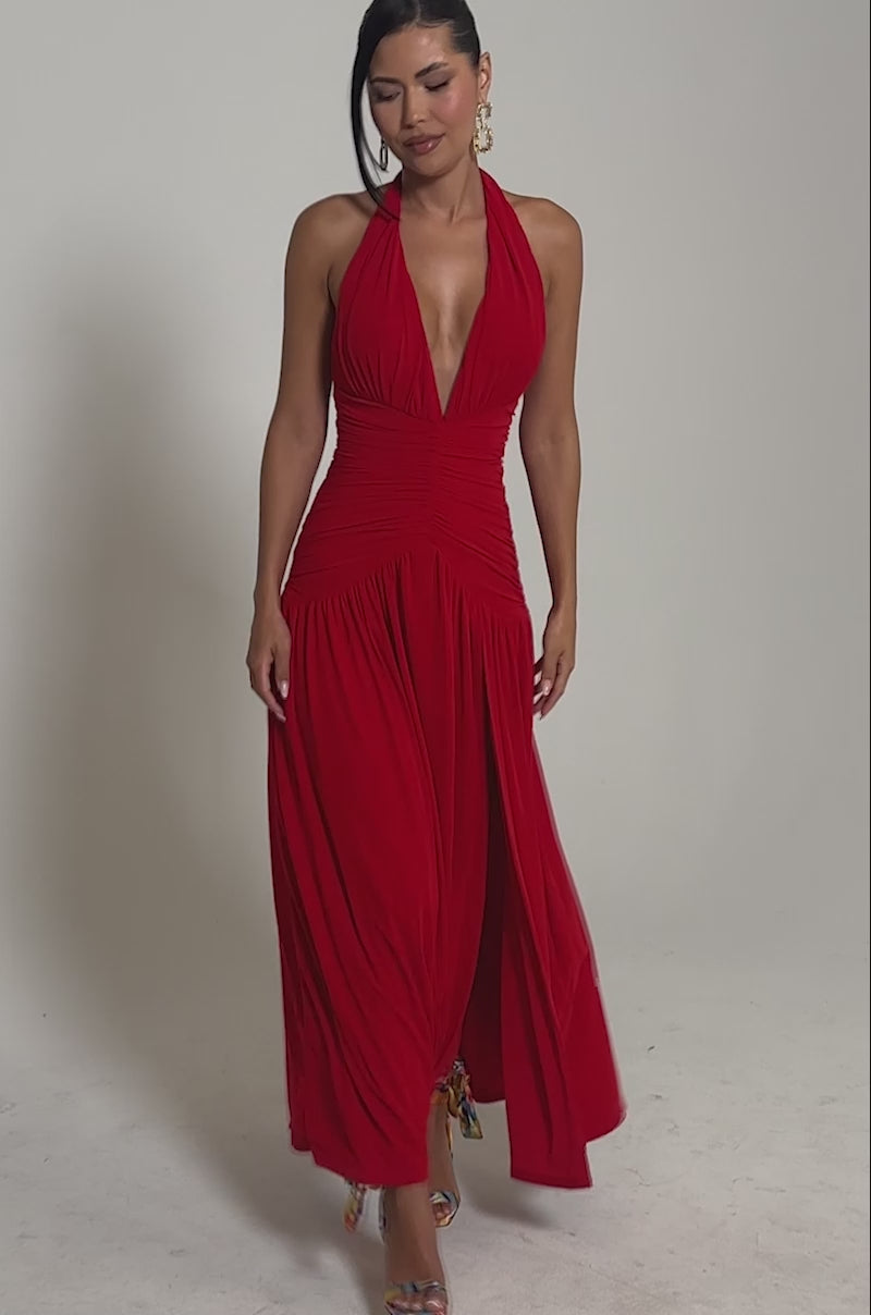 Video of woman modeling the SUGAR RUSH MESH MAXI DRESS, a red ruched halter maxi with a high slit and plunging neckline. She pairs it with colorful heels, gold earrings, and a side bun against a plain backdrop.