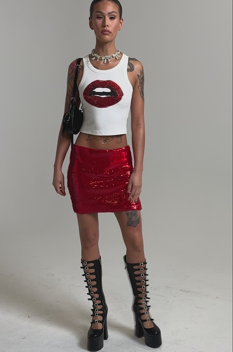 Video of woman modeling the KISS ME BABY TANK, a cropped graphic racerback featuring red sequin lips, paired with a matching red sequin mini skirt. She wears a necklace, has slicked-back hair, and stands against a plain background.