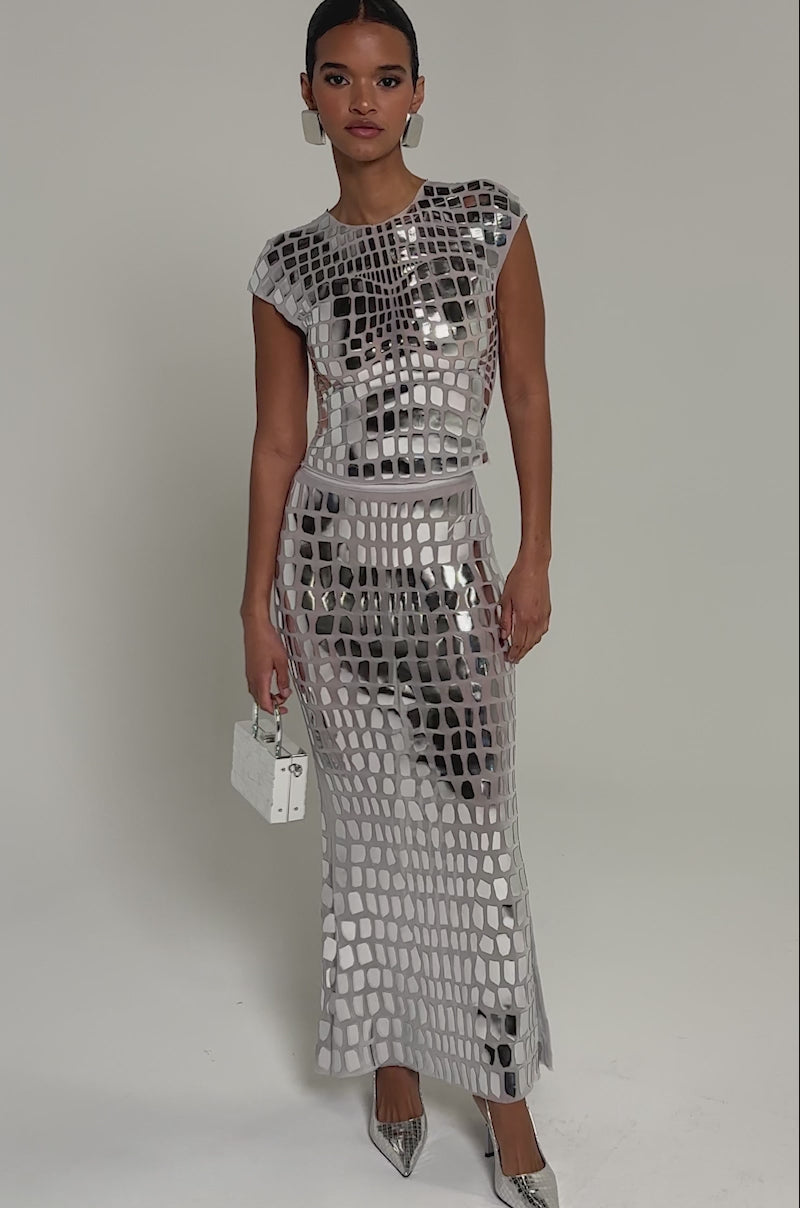 Video of woman modeling the CHAMPAGNE POPPIN MAXI SKIRT, a fitted, floor-length silver skirt with a high-shine, mosaic-like pattern. She pairs it with a small silver handbag and matching pointed-toe heels.