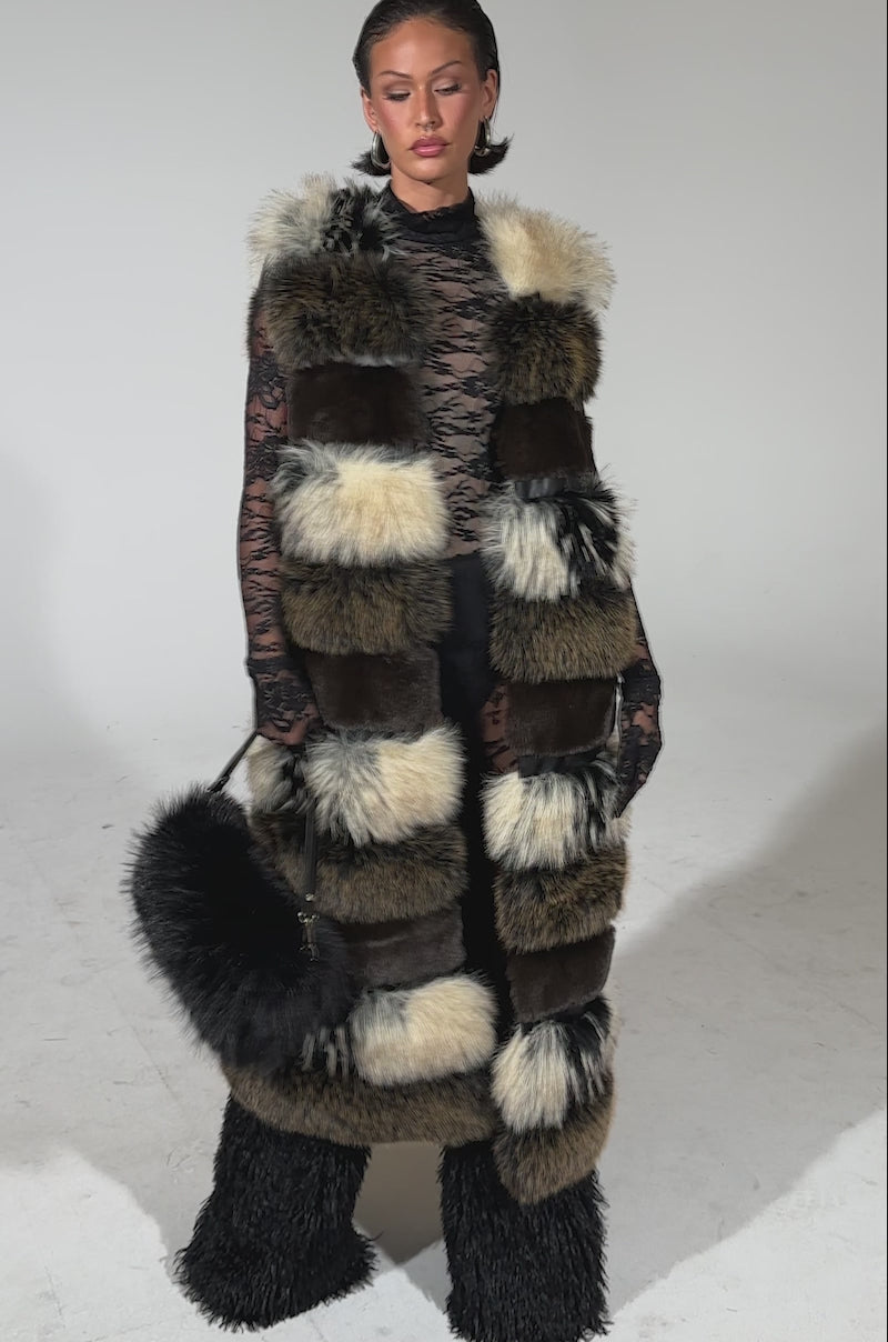 A video of a woman wearing the PORTIA PANELED FUR VEST with brown, black, and cream patches over black lace sleeves and voluminous black furry pants. She carries a small black furry bag and has slicked-back hair.
