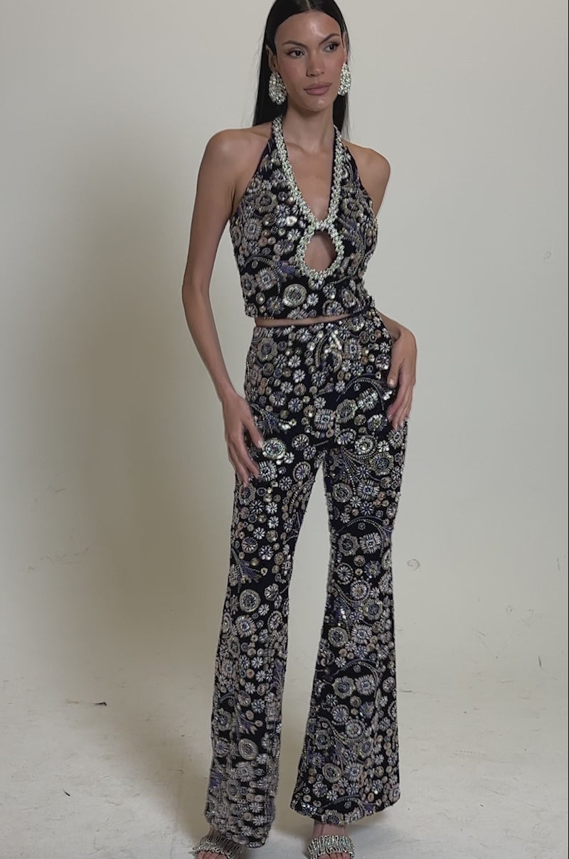 A video of a woman wearing the QUEEN OF EVERYTHING RHINESTONE HALTER BLOUSE in navy blue with matching flared pants, gold and white floral pattern, accessorized with a round metallic bag and high heels, standing against a plain gray background.