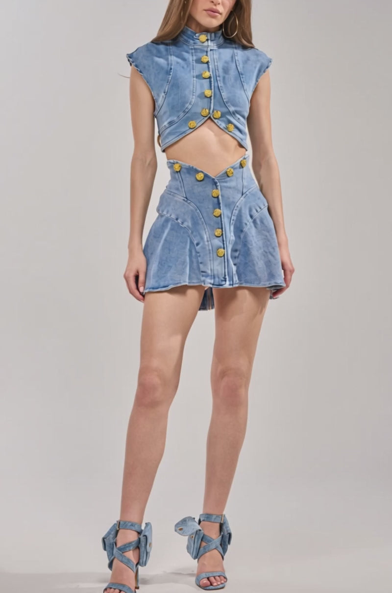 Video of woman modeling the GIVE IT TO ME BABY DENIM MINI SKIRT, paired with blue strappy heels featuring bows against a plain, light-colored background.