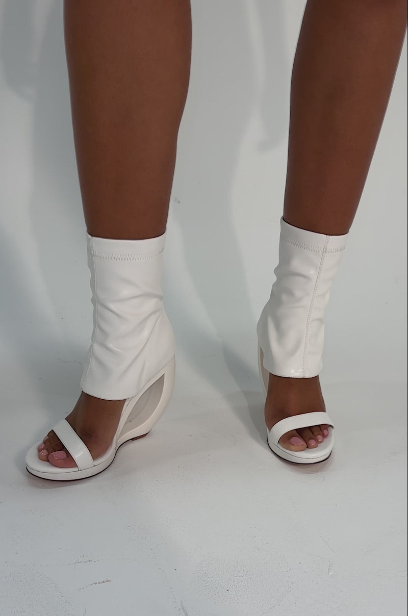 Video of woman modeling the AKIRA GOLD DUMISANI WHITE WEDGE OPEN TOE BOOTIE—an ankle-high, sock-like faux leather bootie with open toes, cut-out futuristic wedge heels, and a zipper—standing out against a plain light background.