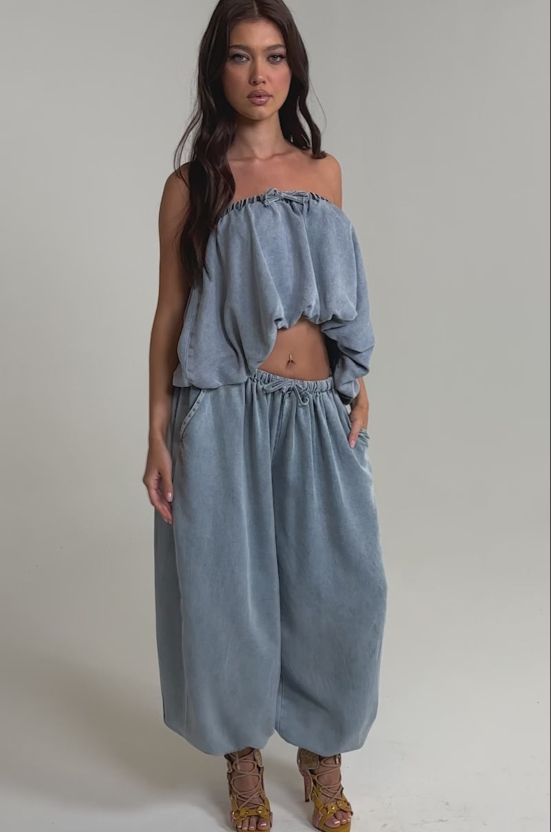Video of woman modeling the PUMPED UP DENIM BLOUSE, a loose, light blue strapless denim crop top with a gathered neckline and matching baggy pants, standing against a plain light background for a relaxed yet stylish look.