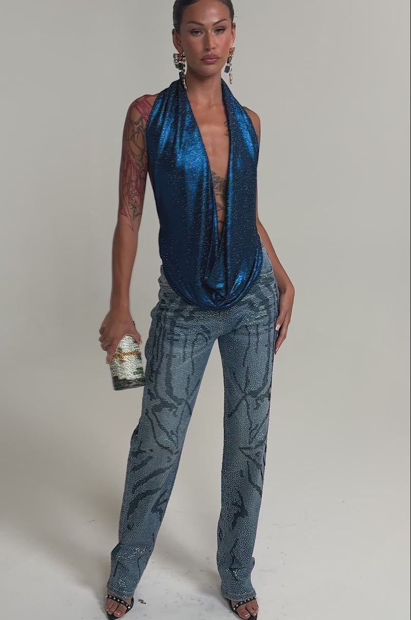Video of woman modeling the MERMAID DREAM RHINESTONE OPEN BACK HALTER TOP, paired with blue jeans and colorful dangling earrings, standing against a neutral background.