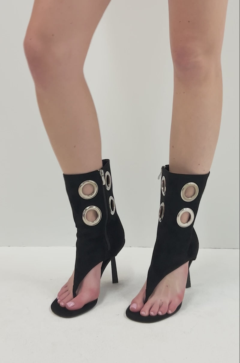 Video of person modeling the AZALEA WANG BAINA BLACK GROMMET DETAIL GLADIATOR SANDAL. The shoe is a black high-heeled sandal with an open toe, high ankle cuff, four metallic grommets on the ankle strap, and a sleek stiletto heel for a bold, modern style.