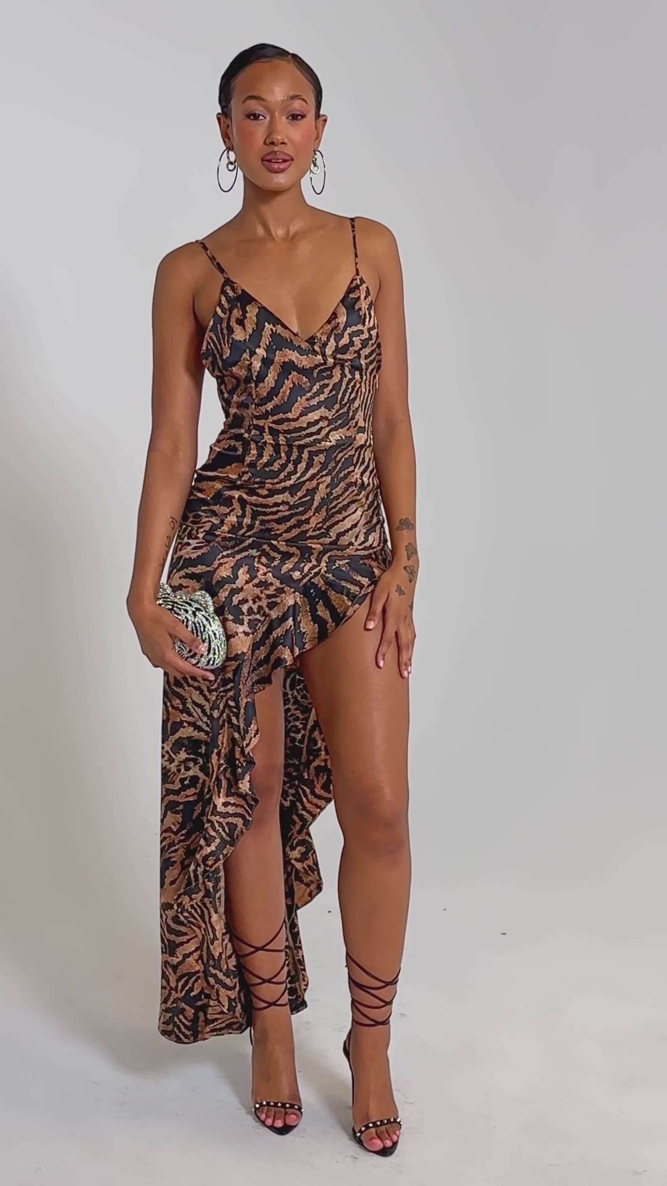 A video of a woman wearing the IN THE WILD ASYMMETRICAL MAXI DRESS—a sleeveless, leopard print maxi with spaghetti straps and high slit—paired with a small clutch and strappy high heels.