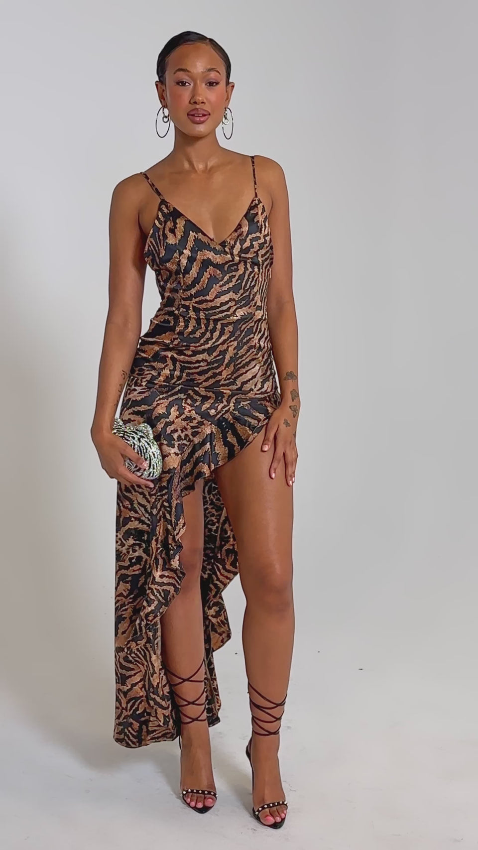 A video of a woman wearing the IN THE WILD ASYMMETRICAL MAXI DRESS—a sleeveless, leopard print maxi with spaghetti straps and high slit—paired with a small clutch and strappy high heels.