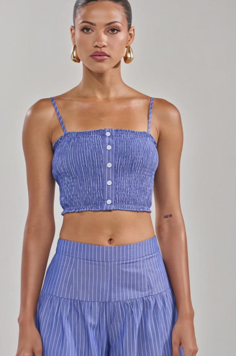 Video of woman modeling the BUSINESS ONLY BRALETTE, a blue pinstriped cotton tank crop top with thin straps and front buttons, paired with gold hoop earrings, and her hair tied back.