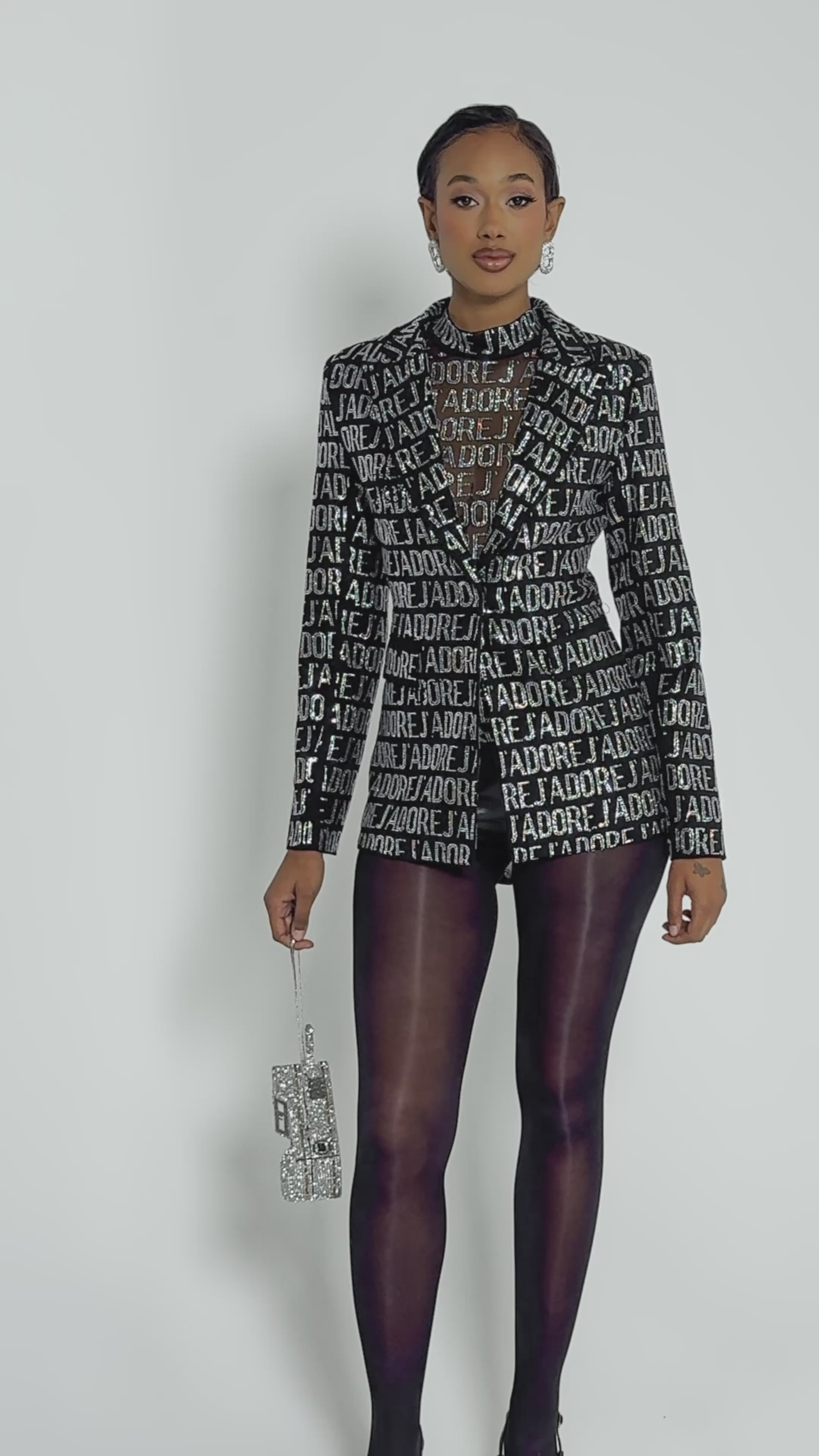 Video of woman modeling the J'ADORE EMBELLISHED BLAZER, adorned with metallic letters, layered over a sheer black top and black tights, set against a plain light background.