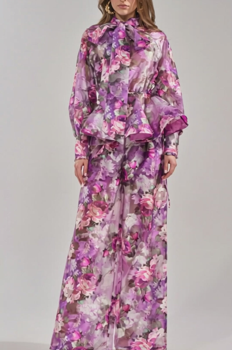Video of woman modeling the KEEP IT FRESH PALAZZO PANT, a sheer, purple floral wide-leg style. She pairs it with a peplum top featuring long sleeves and a bow neck, accessorized with gold earrings and sandals for a resort-ready look.