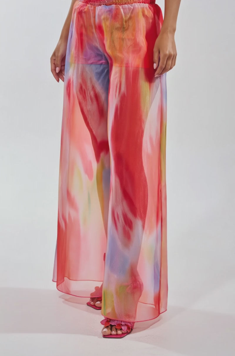 Video of woman modeling the INTERESTED IN YOU PALAZZO PANT, featuring sheer organza with a watercolor print in pink, red, orange, and blue, a red smocked waistband, wide-leg silhouette, and open-toe sandals.