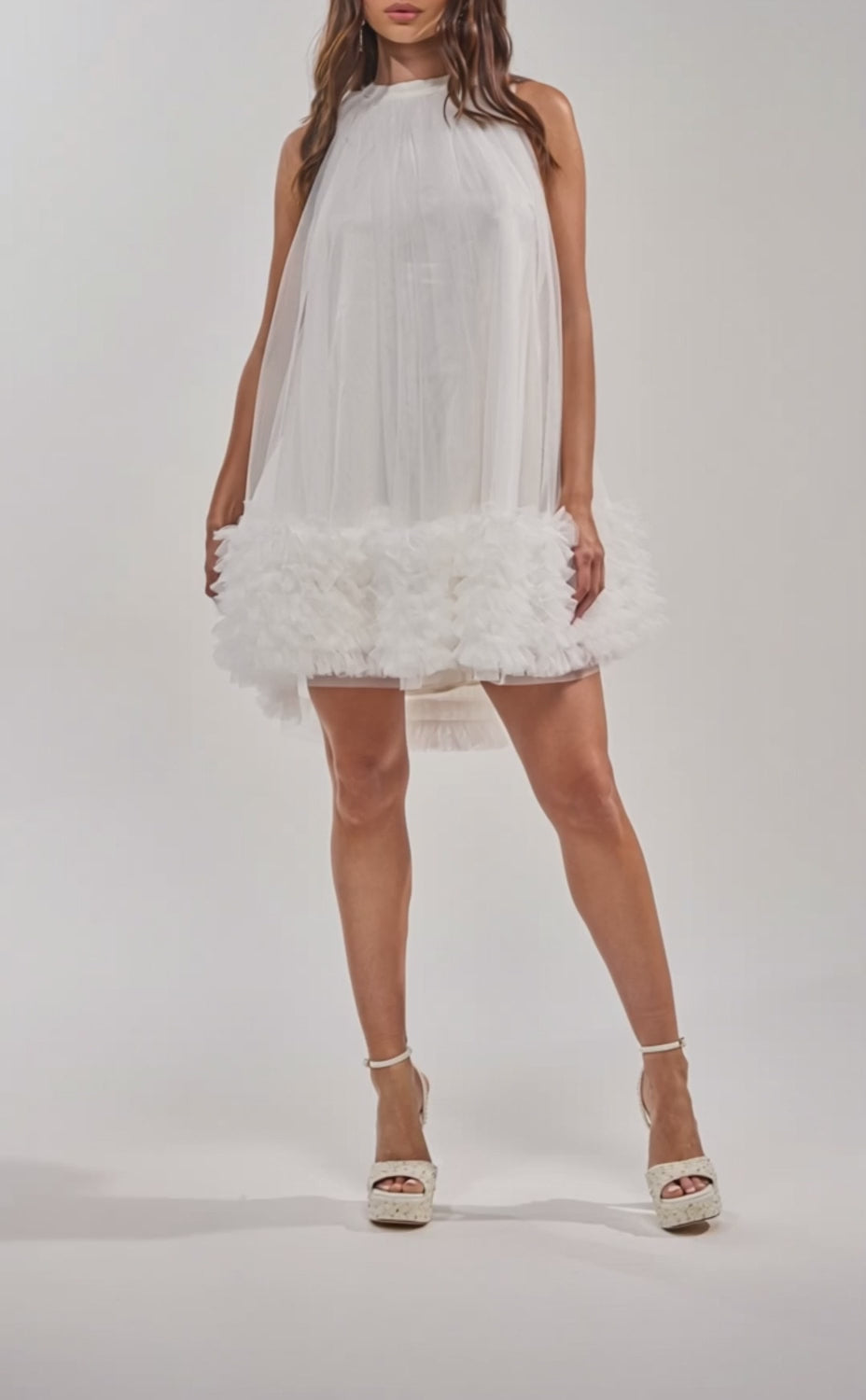A video of a woman in the ARI TULLE RUFFLE MINI DRESS IN WHITE, featuring a sheer overlay and ruffled hem, styled with platform sandals. 