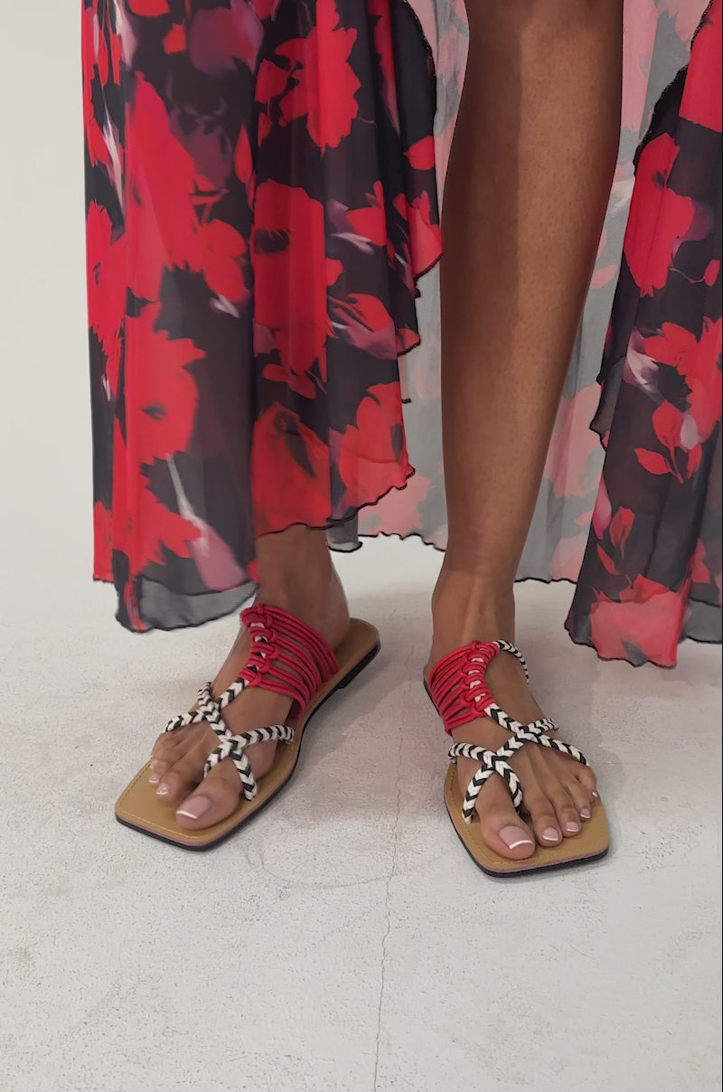 A video of a person wearing a flowy semi-sheer floral skirt and AKIRA SANSANVI RED MULTI ROPE FLAT SANDALS featuring braided red and black-and-white rope straps.
