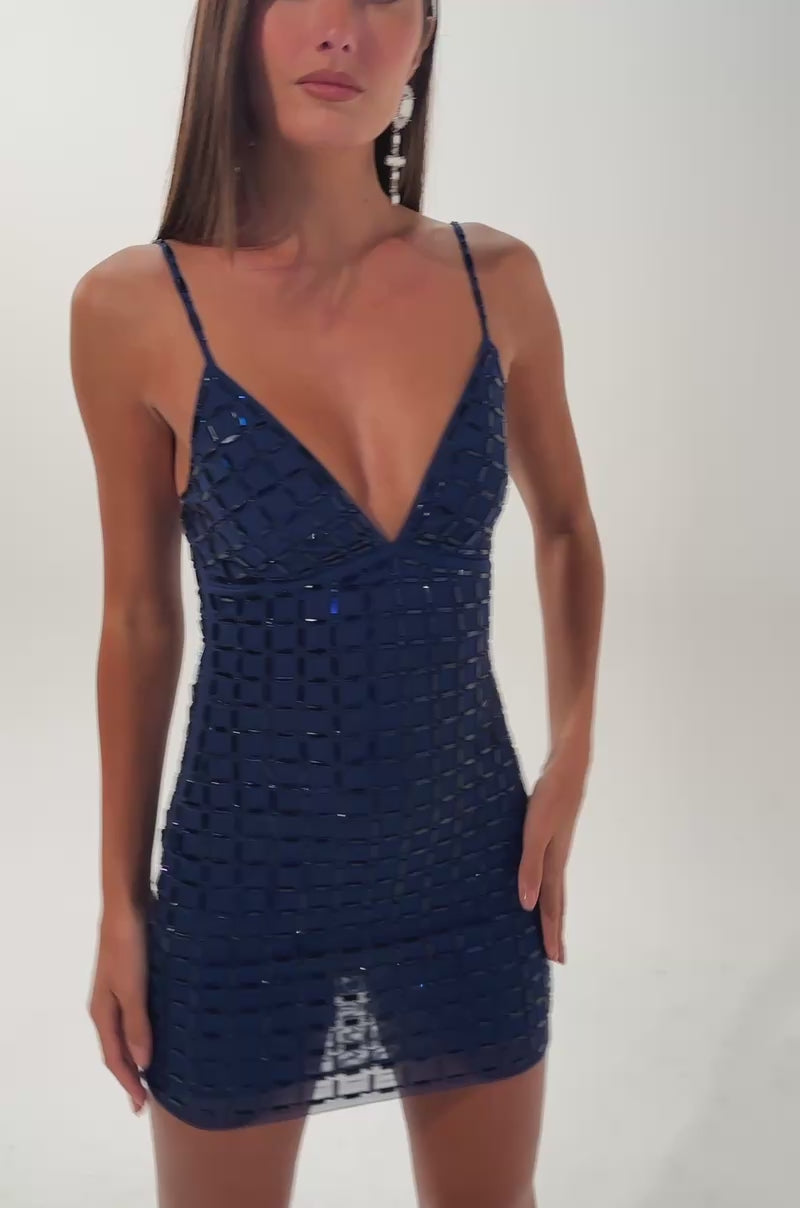 A video of a woman wearing the CITY LIGHTS RHINESTONE MINI DRESS IN BLUE, a sleeveless navy mini with deep V-neck and geometric square pattern, styled for a club look with an ornate clutch purse and statement earrings.