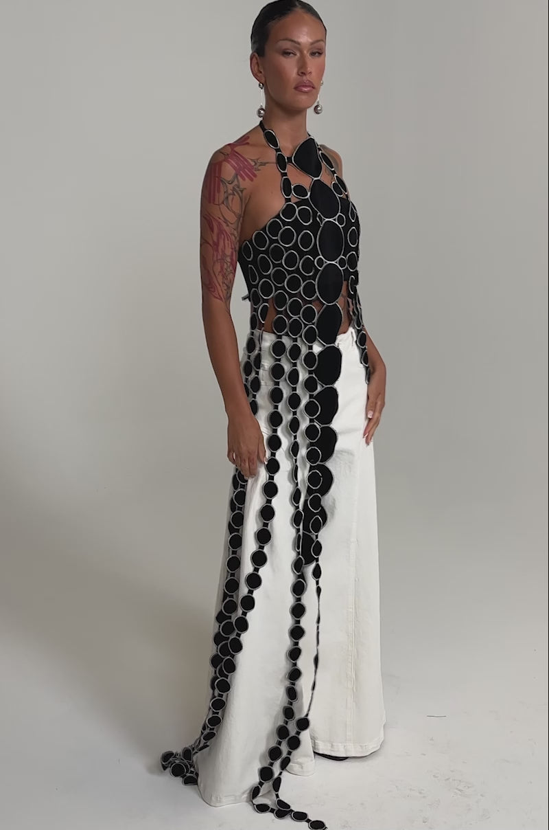 A video of a woman wearing the DISILLUSIONED HALTER TOP, a black and white halter-neck design with cutout details of large black circles connected by smaller ones in vertical rows.
