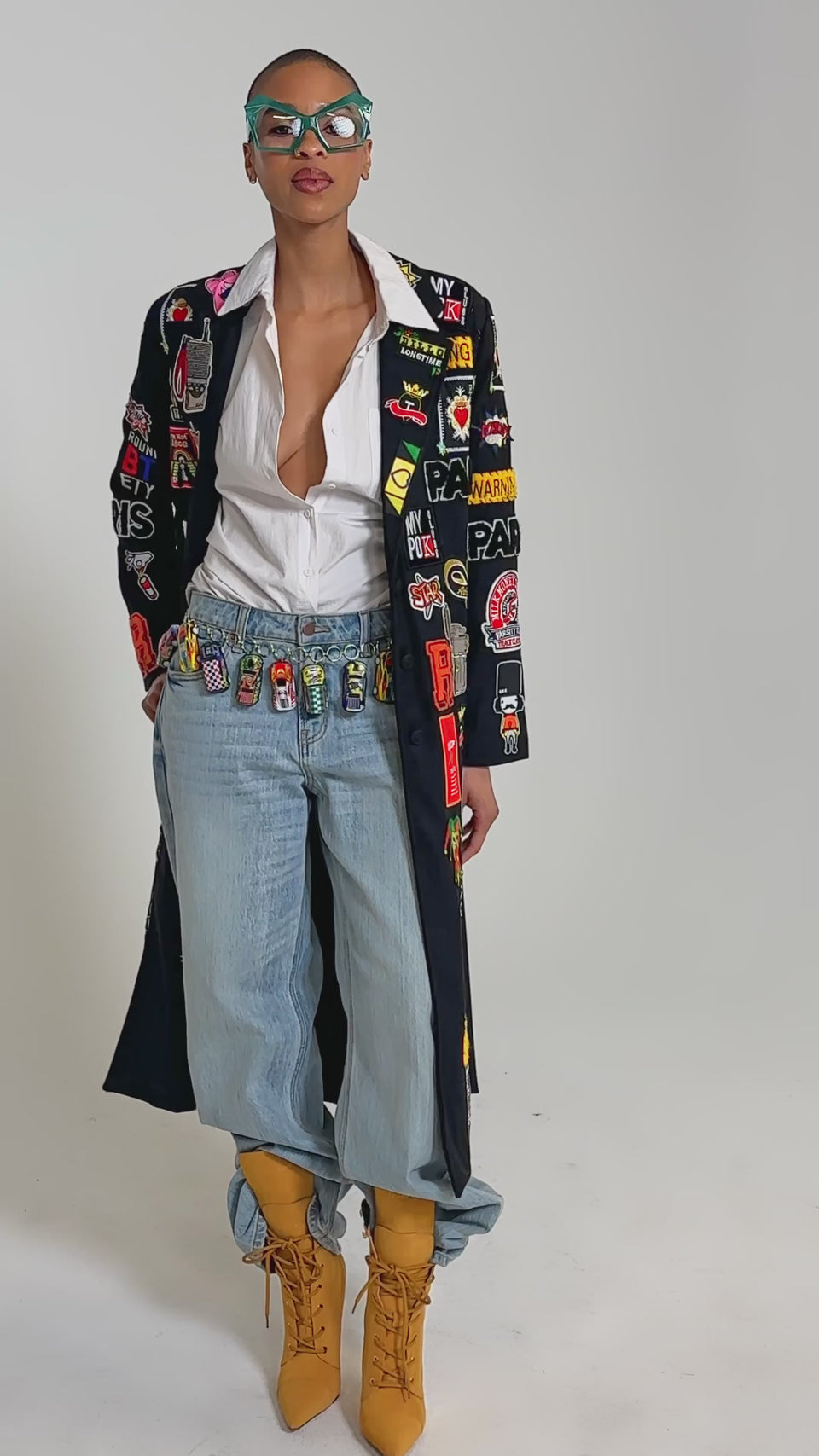 Video of woman modeling the EVERYWHERE I AM PATCHWORK TRENCH over a white shirt, rolled-up blue jeans, and tan high-heeled lace-up boots stands against a plain background.