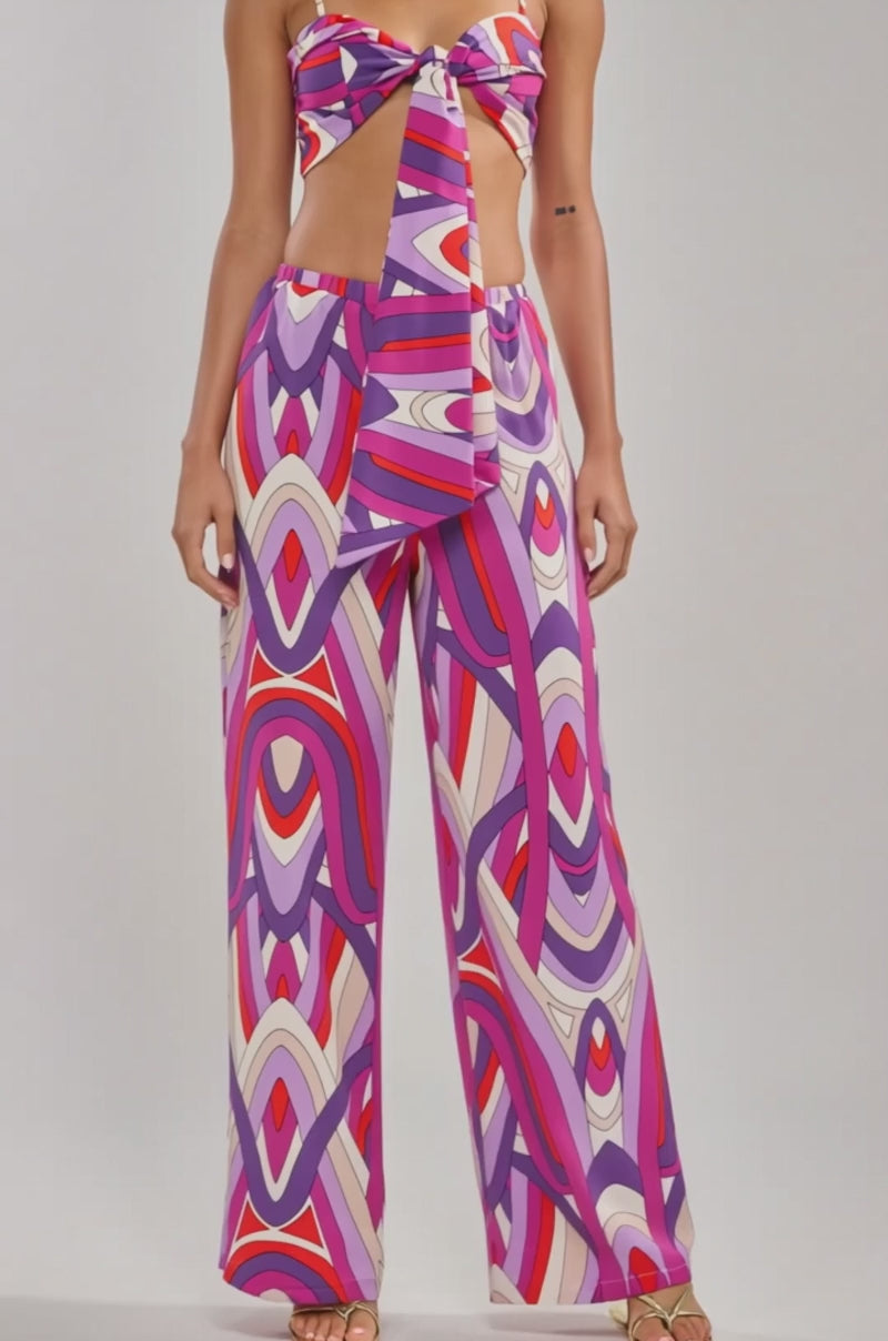 A video of a model wearing the GIRLY POP PALAZZO PANT—high-rise, wide-leg pants with a vibrant geometric pattern in purple, pink, red, and white—paired with gold sandals.