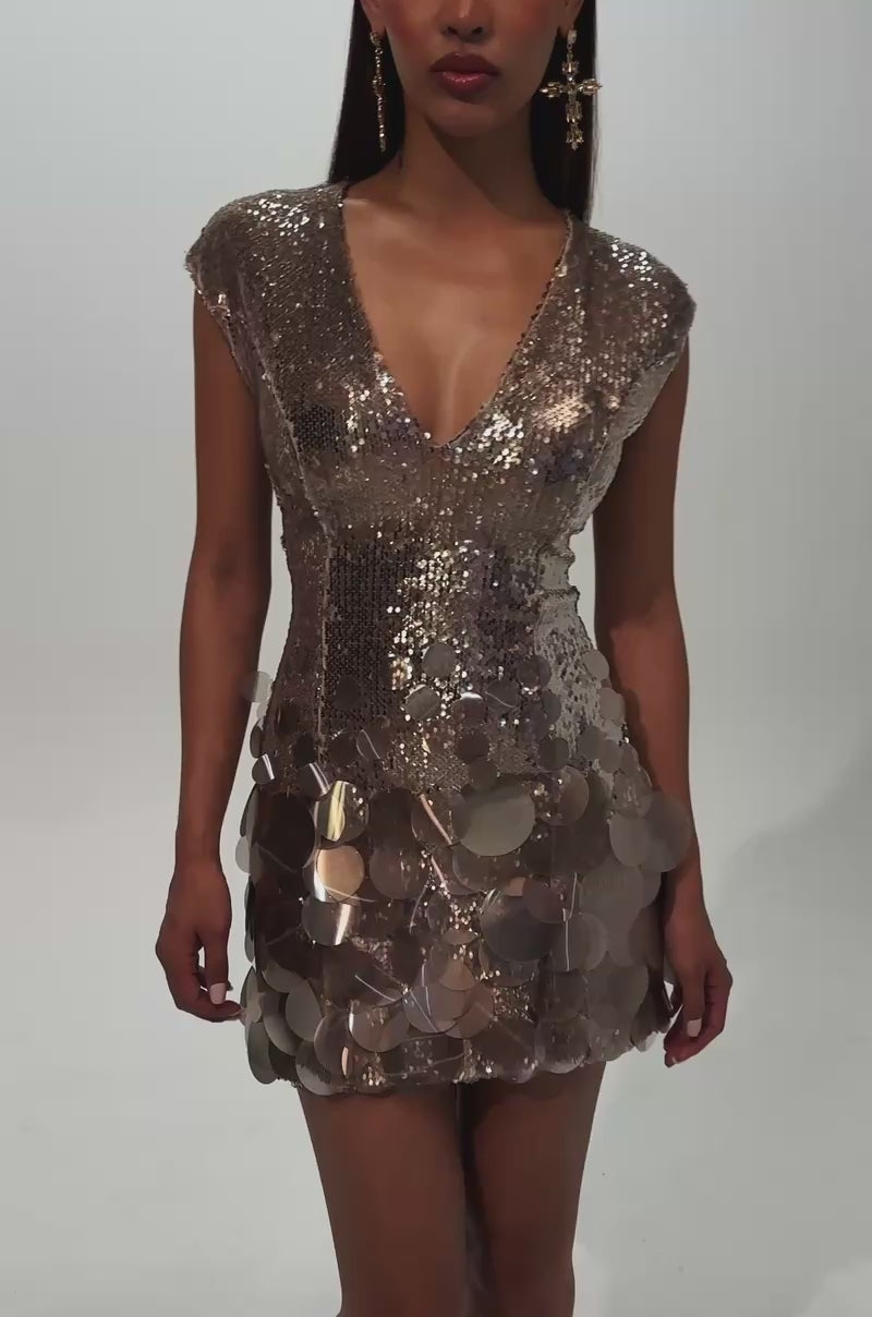 A video of a woman wearing the DANCE THE NIGHT AWAY SEQUIN MINI DRESS IN TAUPE—a glamorous, short party dress featuring gold sequins with large round embellishments and a deep V neckline—paired with statement earrings.