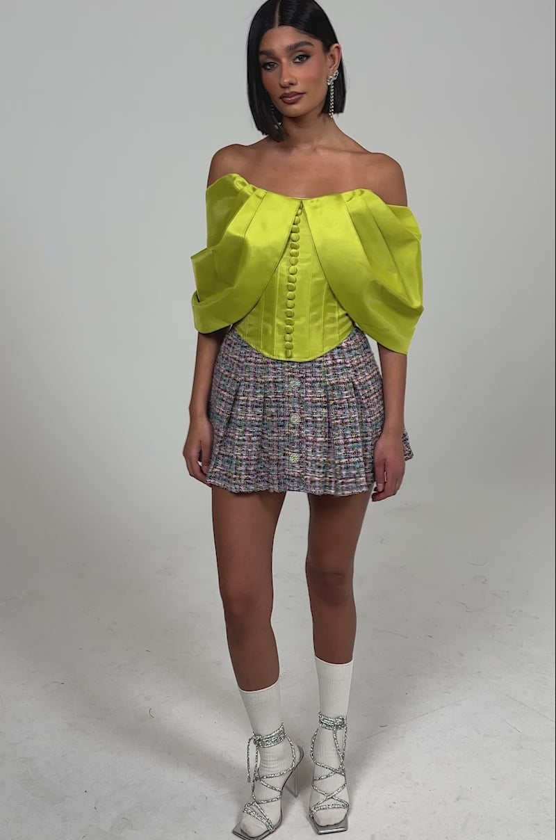 Video of woman modeling the ELECTRIC CORSET IN GREEN styled off-the-shoulder with puff sleeves and front buttons, paired with a high-waisted, multicolored patterned skirt.