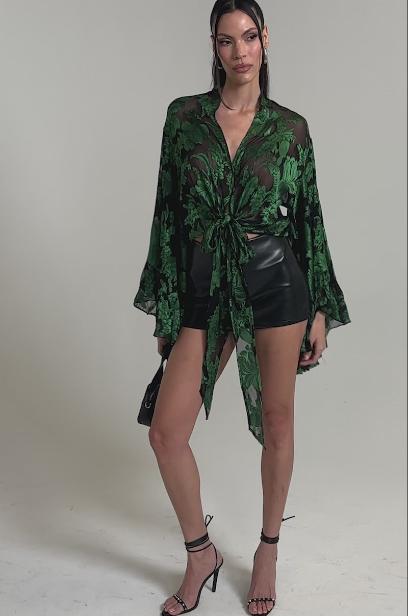 A video of a woman in the IN THE FEELS LACE BLOUSE, a sheer green and black floral crop top tied at the waist, styled with black high-waisted leather shorts and black high-heeled sandals.
