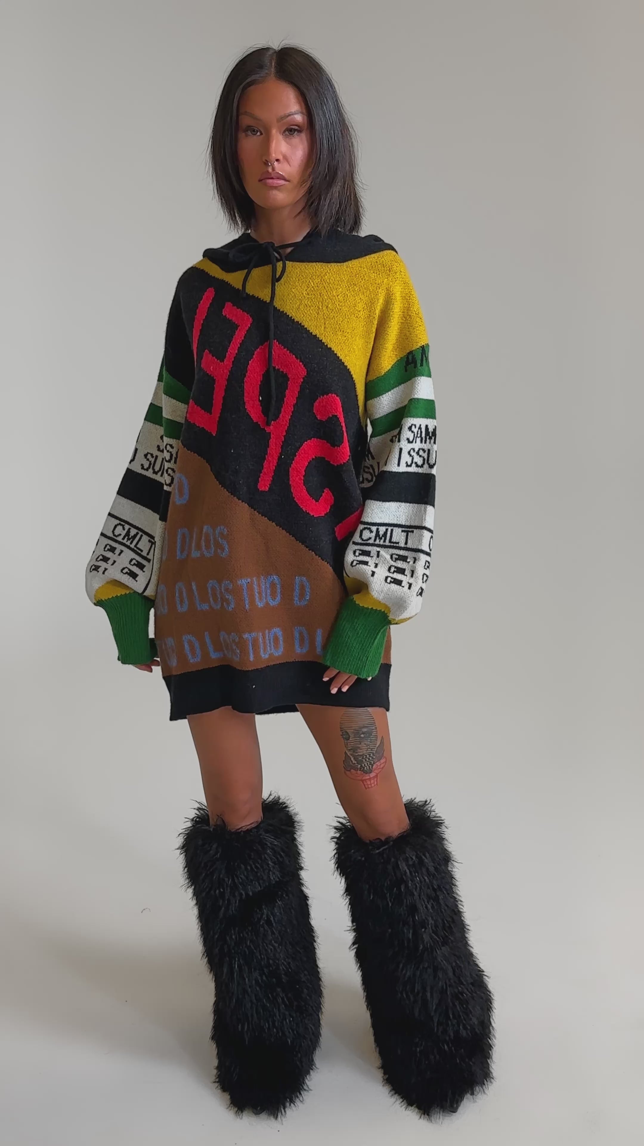 A video of a woman in the ALPHA FEMALE HOODED SWEATSHIRT DRESS IN YELLOW MULTI, a streetwear-inspired, colorful patchwork piece with bold lettering, paired with black furry boots. She has straight dark hair and stands before a plain light backdrop.