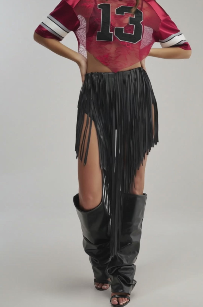 A video of a woman in sunglasses, a red jersey with 13, a COACHELLA FRINGE SKIRT, and tall black slouchy boots stands against a light background for a bold festival-ready look.