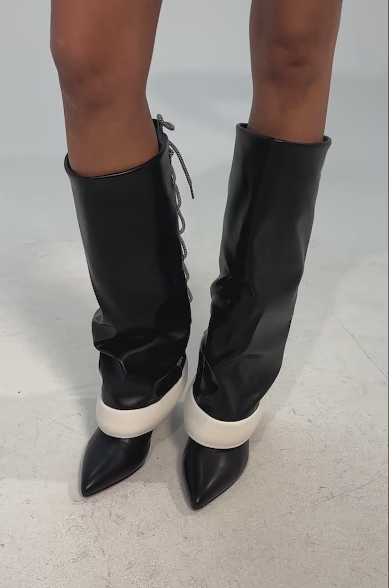 Video of woman modeling the AKIRA GOLD HALOED BLACK LACE UP BACK BOOTIES—knee-high, black and white stiletto heels with pointed toes and crisscrossed gray laces up the back—standing on a light background.