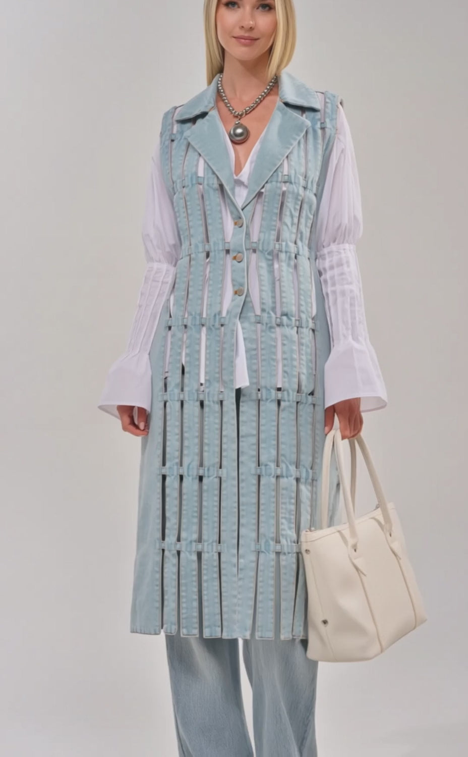 Video of woman modeling the RIGHT THROUGH ME CUTOUT DENIM VEST with vertical cutouts, styled over a white blouse and light blue jeans, accessorized with a white handbag and a chunky silver necklace.