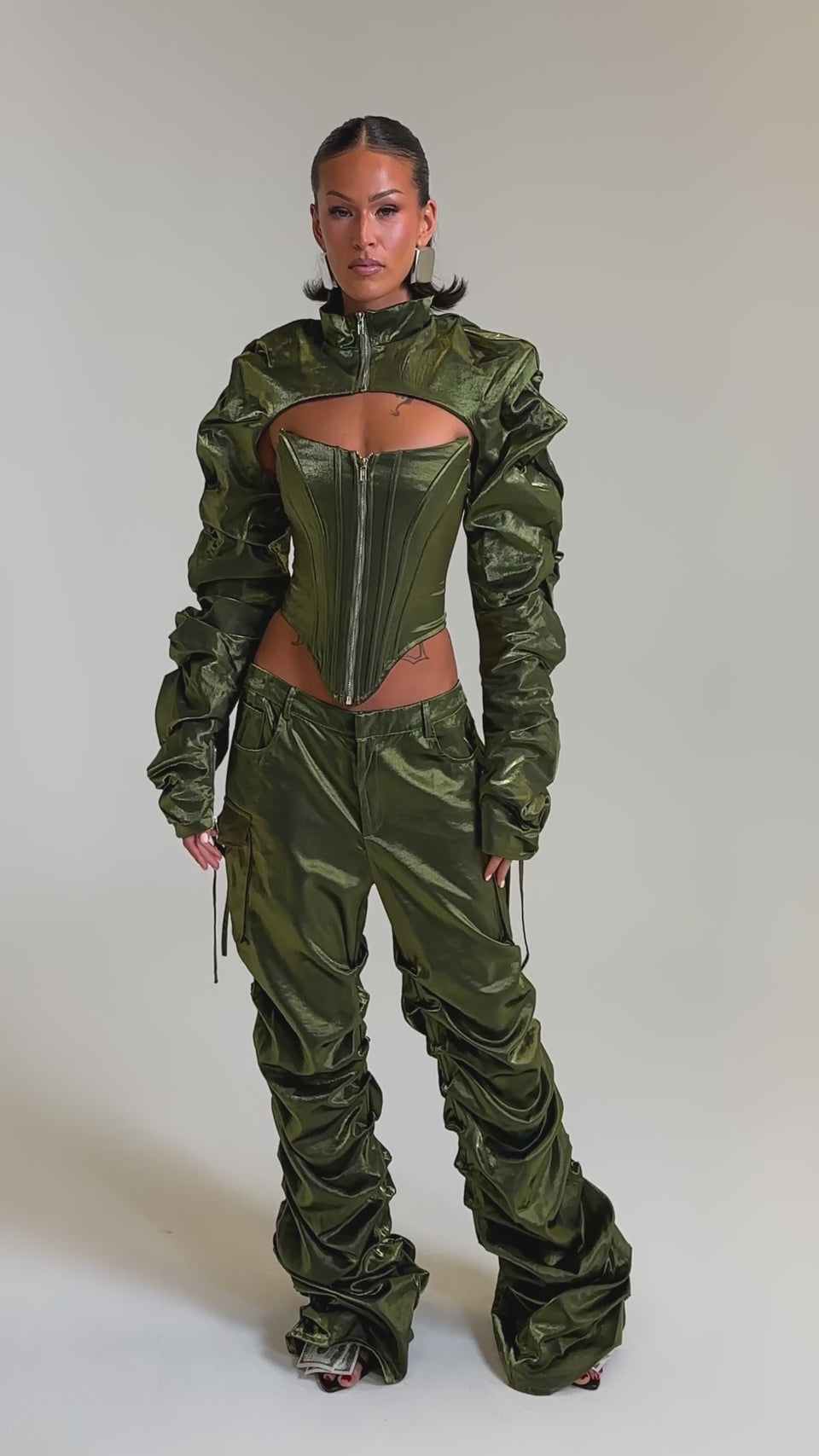 A video of a woman in an olive green, textured pantsuit with ruched details and a zip-up corset top, styled with the LUCKY ONE BOLERO and standout dollar bill print shoes. 