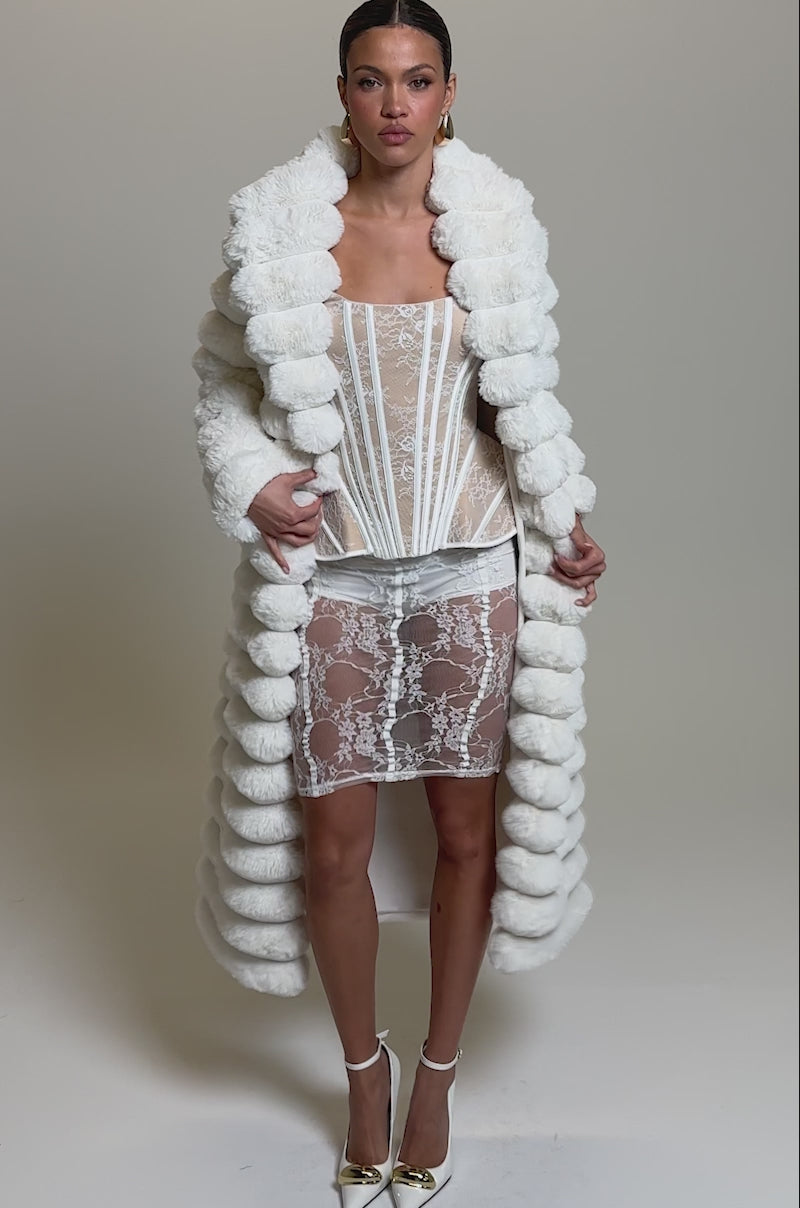 Video of woman modeling the MAXI POPCORN FAUX FUR IN WHITE coat with thick horizontal segments, paired with white heels featuring gold accents and hoop earrings, set against a plain light gray background.