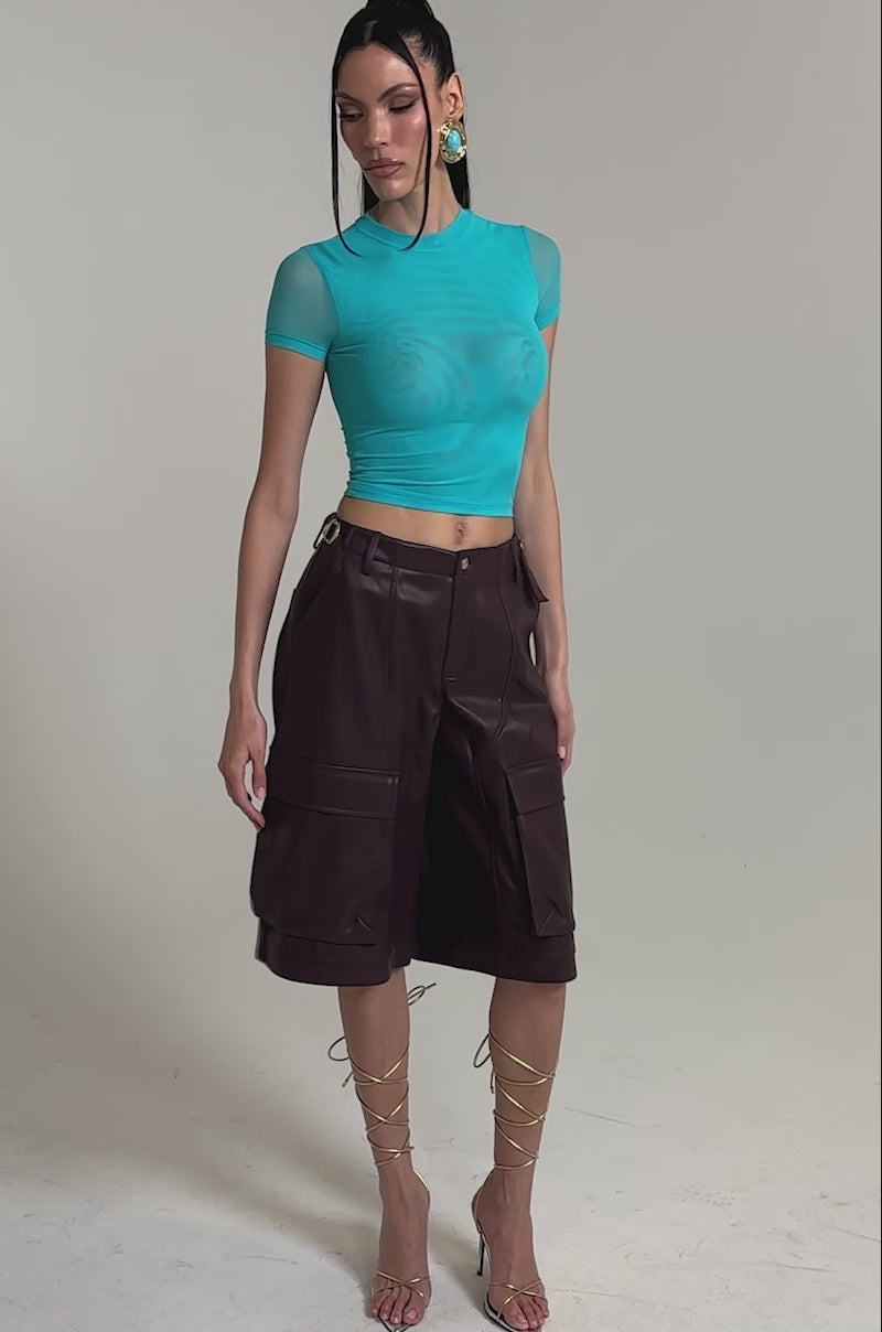 A video of a woman wearing the PAXTON Double Lined Mesh Short Sleeve Tee in turquoise, high-waisted brown knee-length cargo shorts, strappy gold heels, and statement earrings.