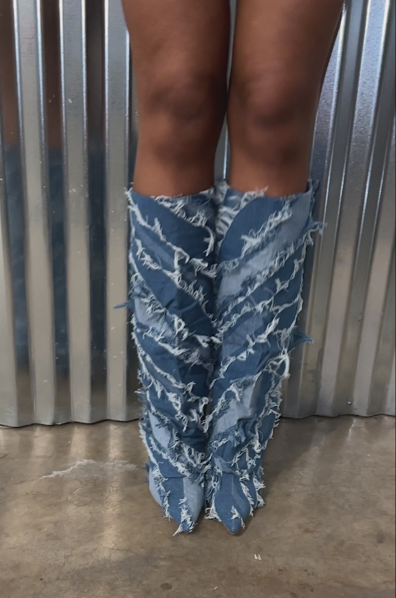 Video of person modeling the AZALEA WANG FISHER DENIM STILETTO BOOT. The boot is a tall, knee-high shaft boot adorned with layered, frayed denim strips, creating a textured and distressed appearance with its blue jean fabric and a thin stiletto heel