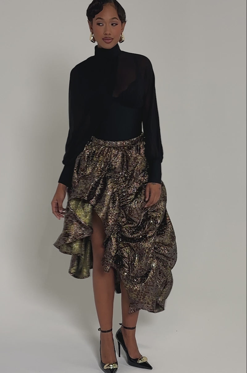 Video of woman modeling the CRAZY FOR YOU BROCADE RUCHED MIDI SKIRT, styled with a black sheer blouse, black and gold heels, and statement earrings against a plain gray background.