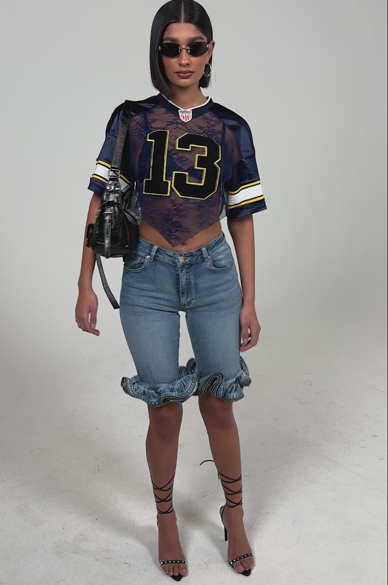 Video of woman modeling the LONDON BERMUDA SHORT, high-waisted denim shorts with a ruffled hem above the knees, styled with strappy black heels and a matching black handbag.