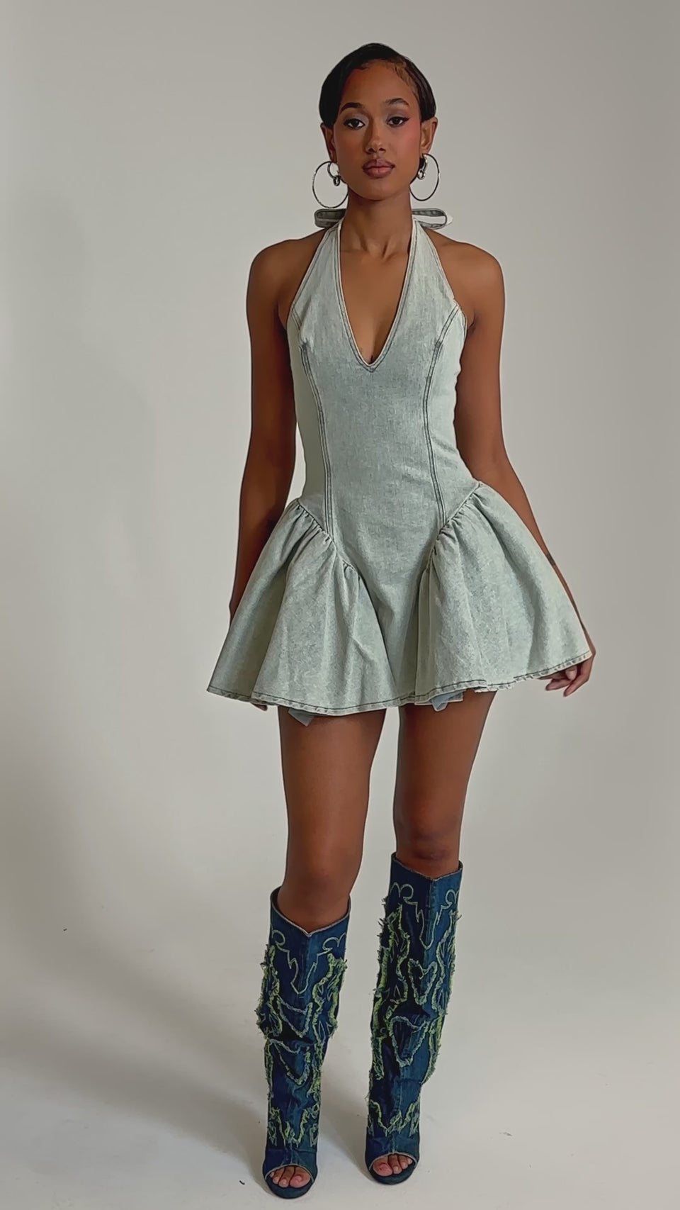 A video of a woman in the STYLE AND GRADE DENIM MINI DRESS, featuring a light blue wash, skater silhouette, and fitted bodice, paired with tall blue boots with green textured designs.
