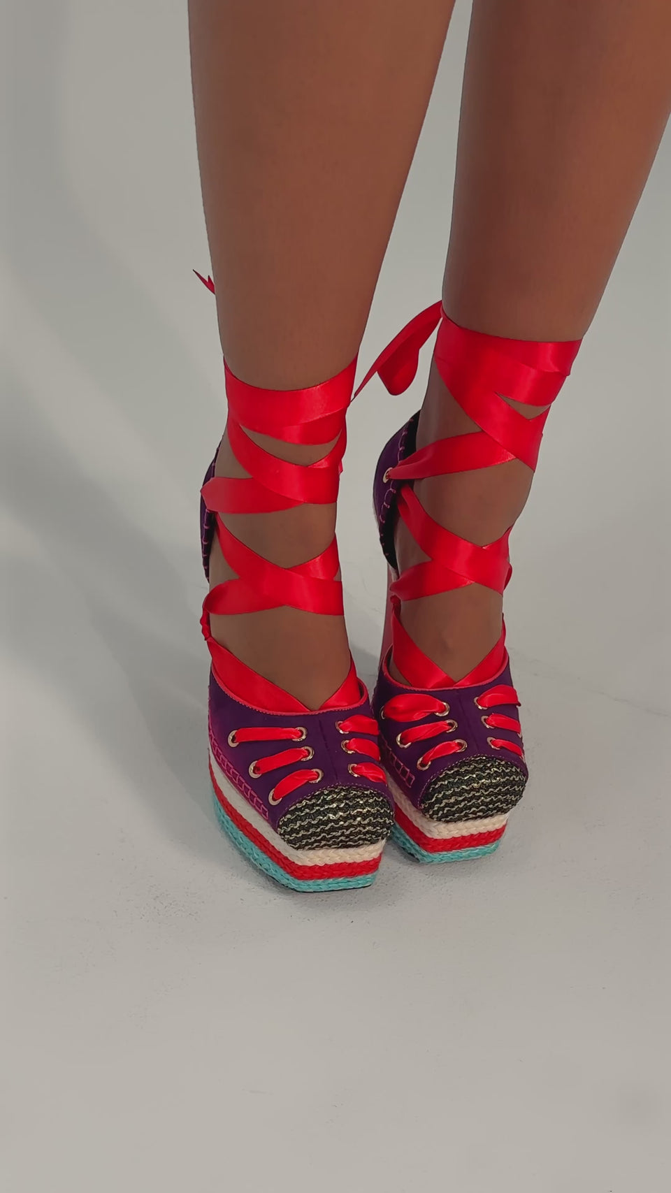 A video of a person wearing the AKIRA GOLD FONTEYN RED MULTI CHUNKY PLATFORM LACE UP PUMP with colorful accents, red ribbon ankle ties, and woven details stands on a light surface.