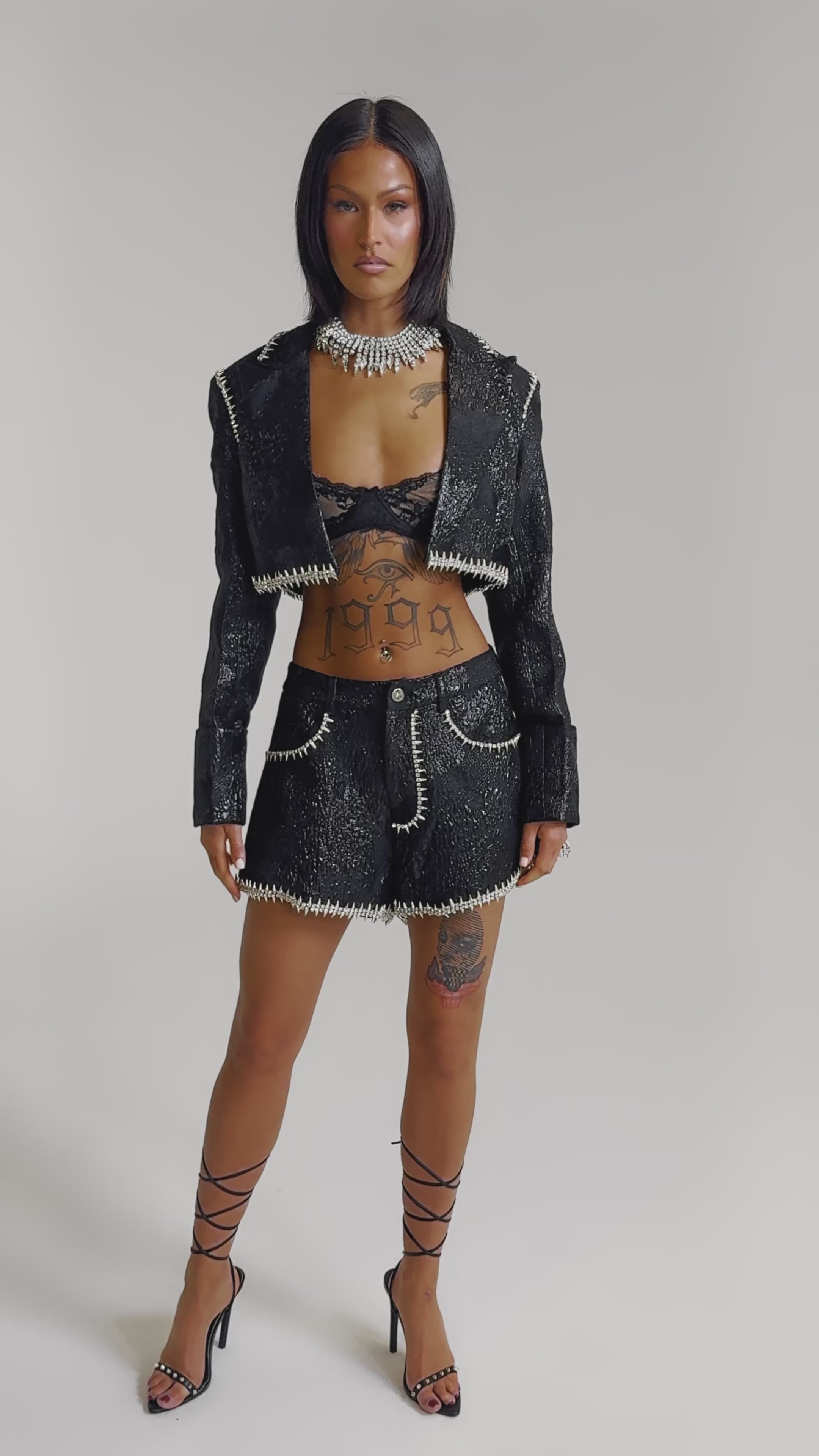 A video of a woman in the TWILIGHT ZONE EMBELLISHED BROCADE SHORTS, a cropped black jacket, lace bralette, and strappy heels. 