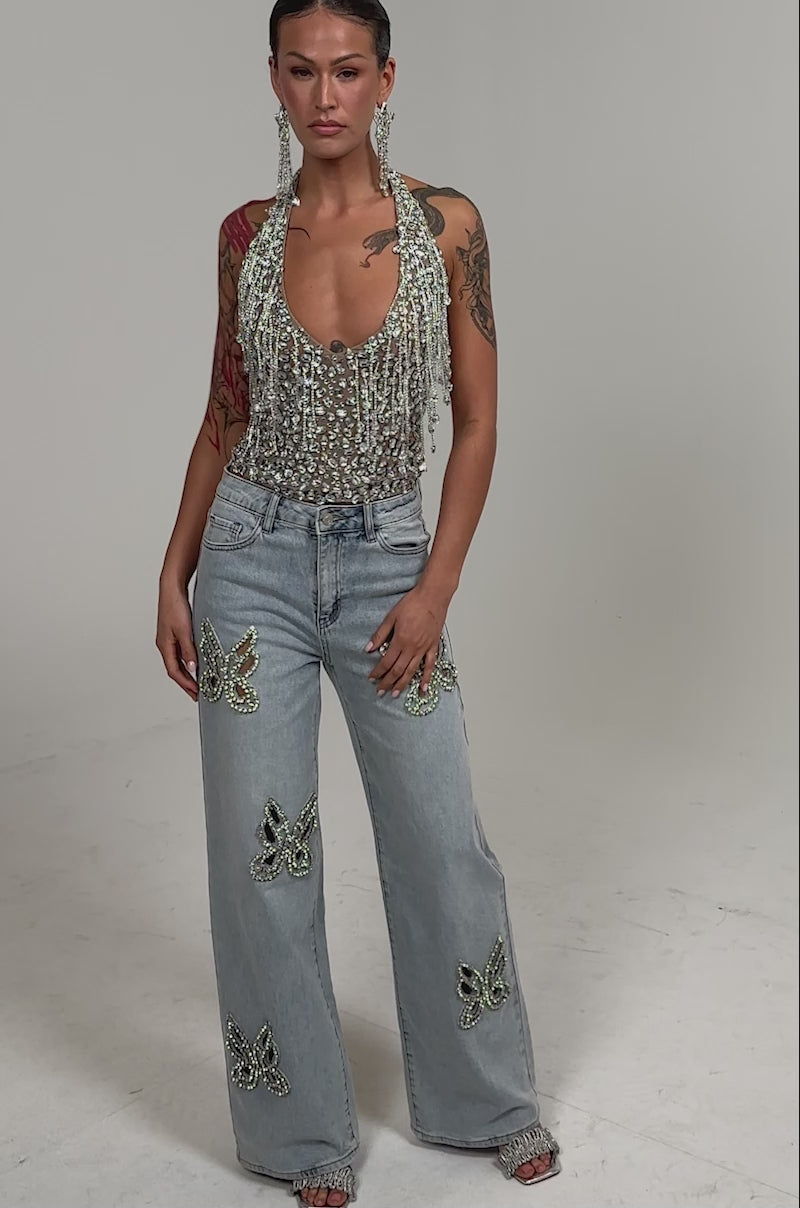 Video of woman modeling the HOTTEST ON THE BLOCK RHINESTONE BLOUSE—a bedazzled halter top with silver sequins and chain fringe—paired with light blue jeans and bold earrings, her tattooed arms visible against a plain background.