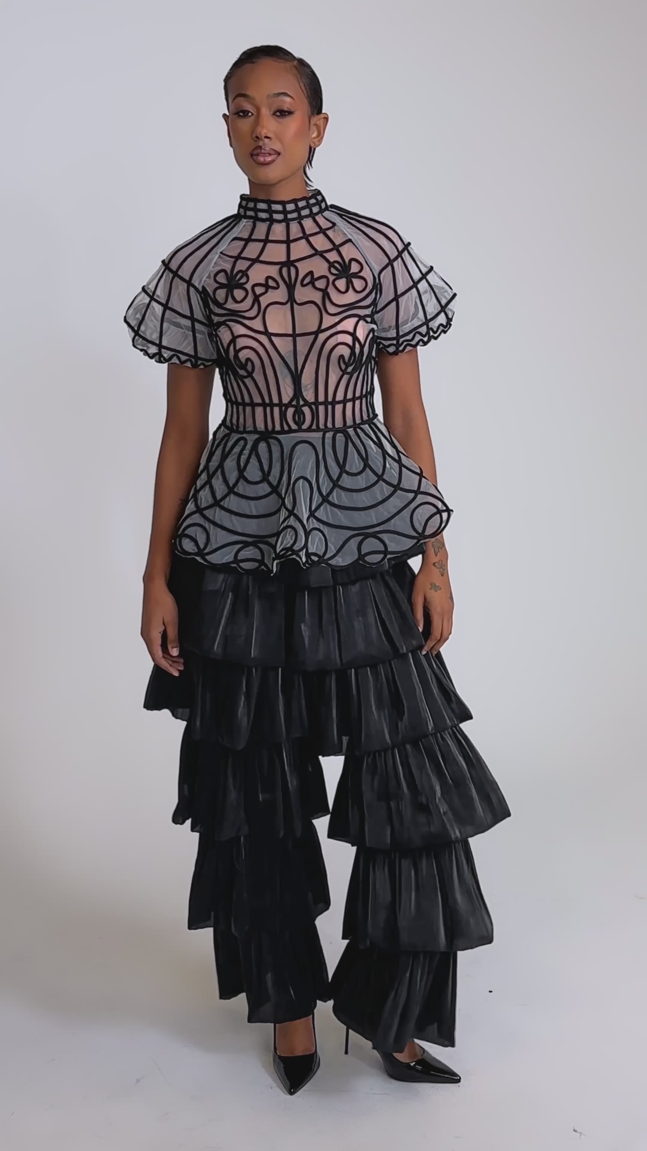 A video of a woman in the DOLL FACE SHORT SLEEVE STRUCTURED BLOUSE, a sheer top with black swirls and ruffled sleeves, paired with tiered black ruffle pants and black heels against a plain background.