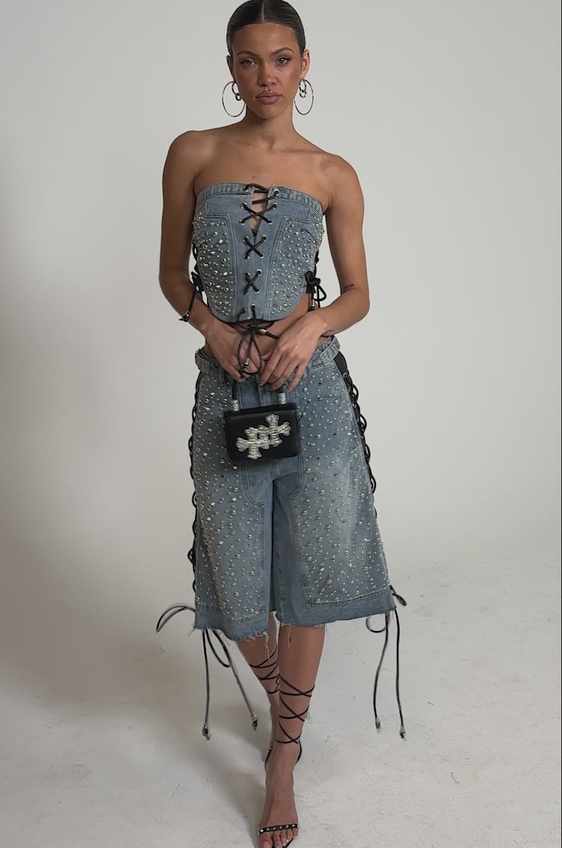 A video of a woman wearing the HONEY BABY LACE UP CORSET with matching studded lace-up shorts, strappy heels, hoop earrings, and carries a small black handbag.