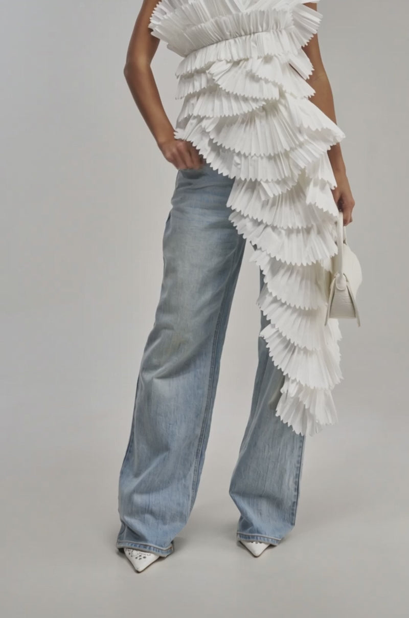 A video of a woman poses in wide-leg light blue jeans and the LUXURIOUS ASYMMETRICAL TOP IN WHITE, featuring dramatic pleats and layered ruffles. 